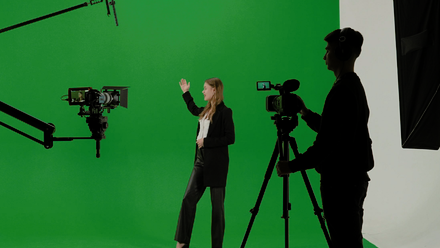 Image of a presenter stood in front of a green screen with cameras and a camera person in silhouette in the foreground.