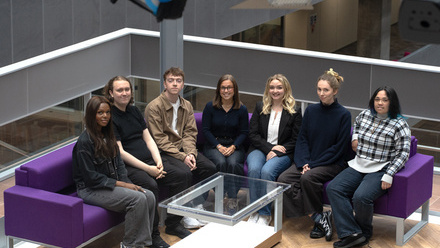 A diverse group of seven young people sit on a deep purple sofa in a light, bright atrium space.