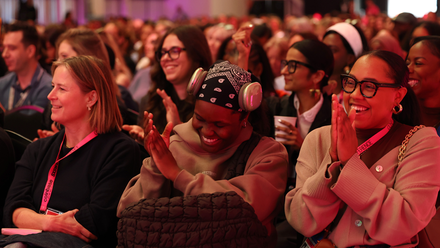 A large, diverse audience seated in rows clap and smile.