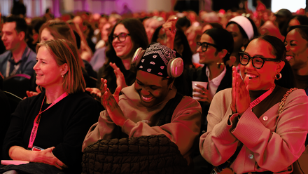 A large, diverse audience seated in rows clap and smile.