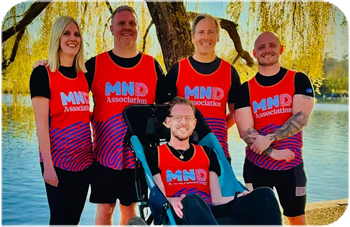 A group of five people in MND running vests pose for the camera in front of a lake. Matt Cox is sat in a mobility buggy in the middle.