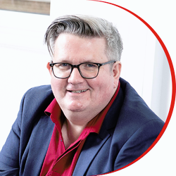 Headshot: Chris Curley, a man with short grey, quiff hair wearing glasses and a dark blue jacket with a red shirt.