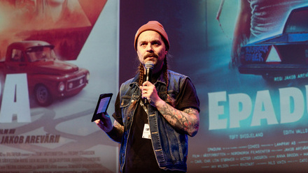 A speaker at Goteborg Film Festival holding a microphone and tablet speaks on stage in front of a large movie poster background.