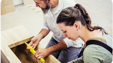 An older man holding a measuring stick and a younger woman in work overalls kneel over an under construction bench. 