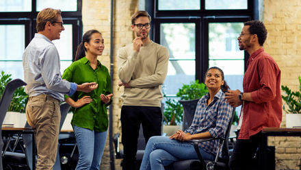 Photo of five diverse colleagues in an office space, they are stood and sat closely together and are engaged in a friendly conversation.