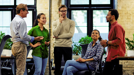 Five diverse colleagues in an office space, they are stood and sat closely together and are engaged in a friendly conversation.