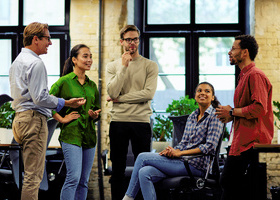 Photo of five diverse colleagues in an office space, they are stood and sat closely together and are engaged in a friendly conversation.
