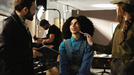 Actor on a film set, sat in a stool having makeup applied whilst another crew member talks to her whilst holding a tablet.