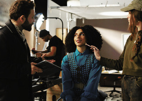 Actor on a film set, sat in a stool having makeup applied whilst another crew member talks to her whilst holding a tablet.
