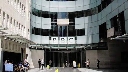 Large glass entrance to BBC Broadcasting House on a bright day with people going in and coming out of the building.