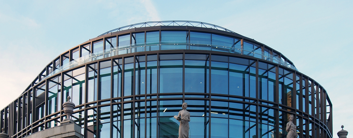 Photo: the top of Channel 4's HQ building in Leeds – a modern glass structure built on top of a traditional stone building with statues. Blue sky above.