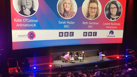 Photograph of a panel of four people on stage in front of a large blue and pink screen in a screening room. 