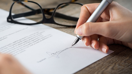 Photo close up of a contract being signed using a silver fountain pen. A pair of glasses are folded and sitting on the desk next to it.