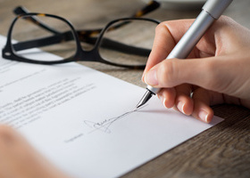 Photo close up of a contract being signed using a silver fountain pen. A pair of glasses are folded and sitting on the desk next to it.
