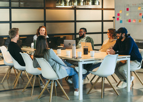 Photo of a diverse team sat around a meeting table in a light office space with a white board full of colourful post-it notes behind them.