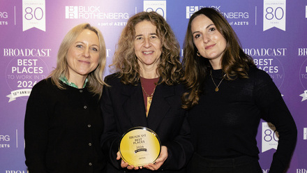 Three women dressed in dark tops holding a circular gold award and stood in front of a purple and blue step and repeat board.