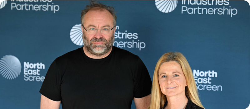 Duncan Gray and Antonia Hurford-Jones, both dressed in black tops, smile for the camera stood in front of a blue North East Screen branding board.