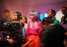 A group of people on a colourful film set, with a camera pointing at them.