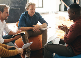 Photo of a diverse group of three men sat in a meeting in an informal part of the office, with notebooks in front of them.