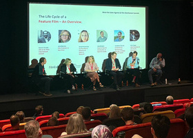 A cinema screening room with a panel of six people sat on stage in front of an audience sat in red cinema seats.
