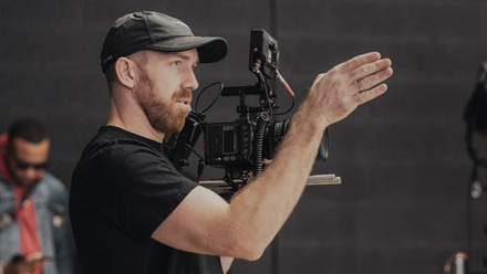 Camera operator wearing a black t-shirt and cap, motioning with his hand.