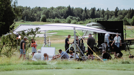 A group of crew members work setting up equipment under the shade of a large sun sail in a large open lawn surrounded by trees.