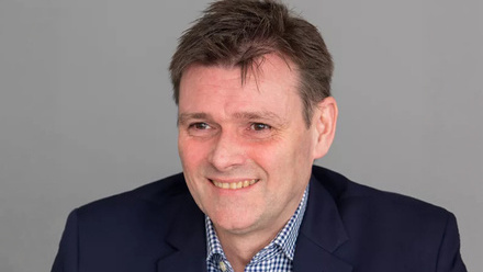 A man with short dark hair wearing a dark blue blazer and light blue shirt.