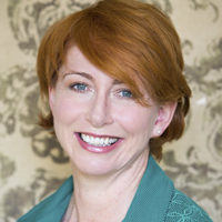 Headshot: Jane Kelly, a white woman with red hair and blue eyes. She is wearing a green jacket and smiling at the camera.