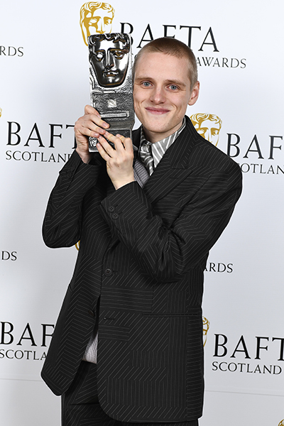 Lewis Gribben, a young white man with very short fair hair and wearing a black suit, holds the BAFTA Scotland Actor Television Award for 'Somewhere Boy'. He is standing in front of branding boards.