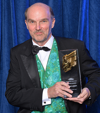 Photo: Stephen Lambert dressed smartly with a green waistcoat and dark suit jacket, holding his RTS Award up, stood in front of a royal blue curtain.