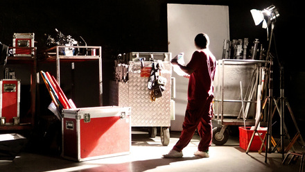 Film crew member standing in a studio among lighting equipment, red cases, and other production gear.