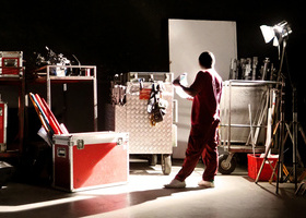 Film crew member standing in a studio among lighting equipment, red cases, and other production gear.