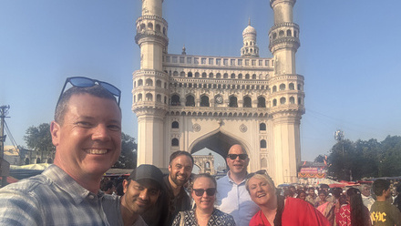 A group of six people pose for the camera in front of an historic Indian archway with clear blue skies above.
