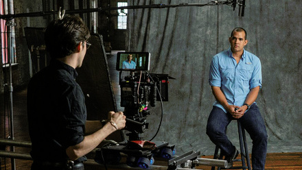 A contributor sits on a stool in front of a neutral backdrop. A large broadcast camera is pointed at him with an operator behind it.