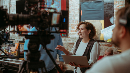 A producer holding a laptop is briefing the crew with a smile on her face, in a brick-walled room with monitors and cameras all around.