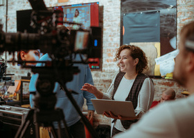 A producer holding a laptop is briefing the crew with a smile on her face, in a brick-walled room with monitors and cameras all around.