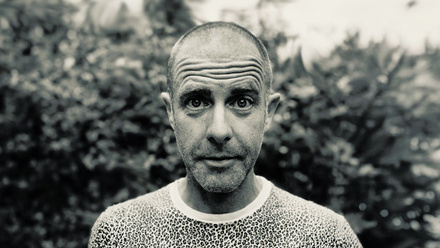 Headshot: Ed Hall, a bald man with a stubble beard and a leopard print top.