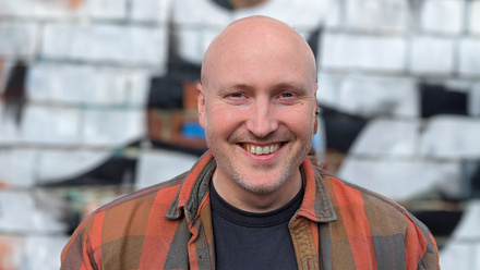 Headshot: Mark Thompson, a bald man with a stubble beard wearing a black t-shirt and an orange checked shirt.