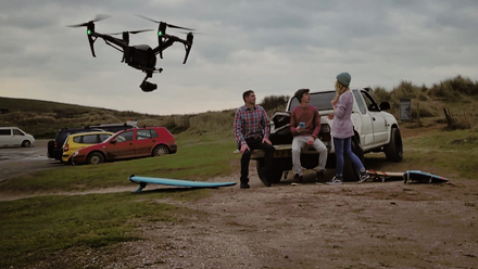 Photo of a large drone hovering in the air with three people sat on the back of a parked pick-up truck in the background, chatting.