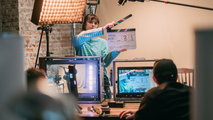 Photo of a woman in a lit studio holding a clapper board up, in the foreground are two men sat looking at monitors on which the woman can be seen.