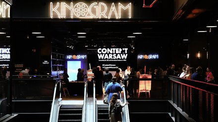 Photo of a large, dark event space with people ascending on an escalator in the centre and the word 'Kinogram' lit up above.