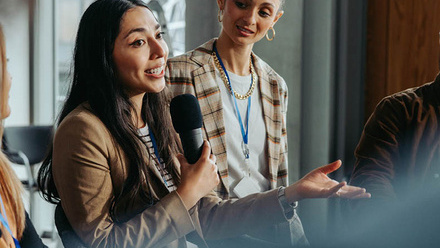 Three people interacting at a conference. One of them is holding a microphone and signalling with their hands.