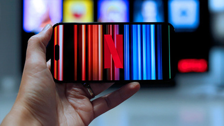 Hand holding a smartphone displaying the Netflix app loading screen in front of a TV displaying Netflix accounts.