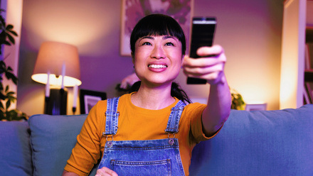 Photo image of a woman dressed casually sitting on her sofa in her living room at night, smiling and holding the remote control up.