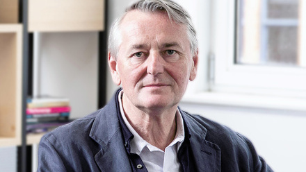 Headshot: Max Rumney, a man with short grey hair. He is wearing a dark blue suit jacket with a light shirt underneath.