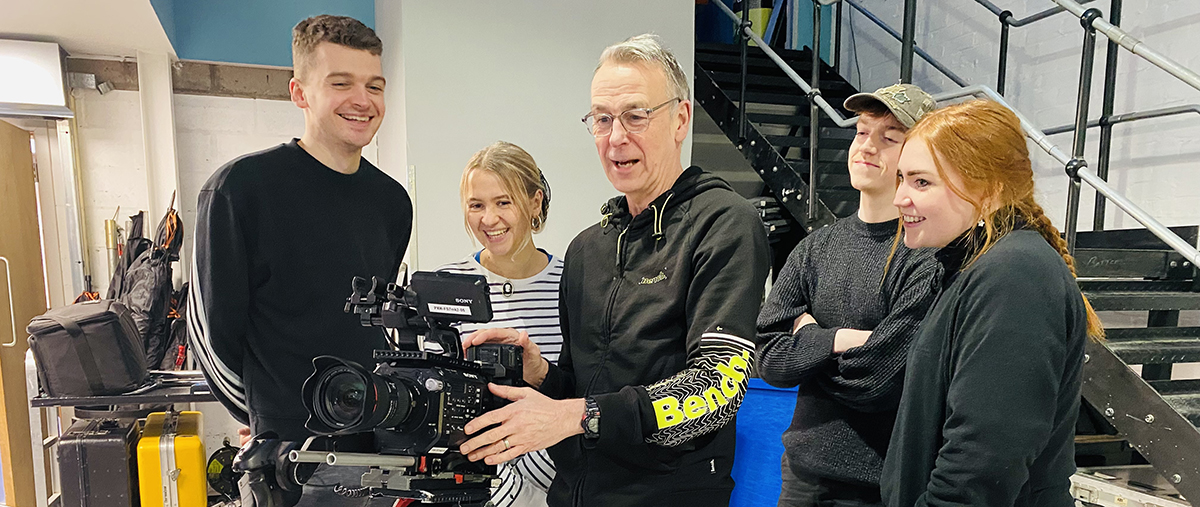 Photo: Four of the 2023 Pact Indie Scheme participants in Scotland are taking part in an NFTS camera training workshop with an expert, who is showing them a camera propped on a tripod.