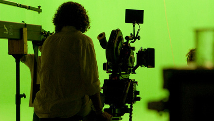 A camera person sitting behind a camera in silhouette in front of a green screen with a subject visible also in silhouette.