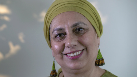 Photo of a woman wearing a lime green headscarf, green earrings and a green top stood in front of a light wall with dappled sunlight on it.