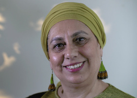 Photo of a woman wearing a lime green headscarf, green earrings and a green top stood in front of a light wall with dappled sunlight on it.