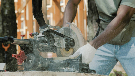 Photo close up of a bench sawing machine being operated by a man with white gloves on, with sawdust flying. A wooden structure is in the background.
